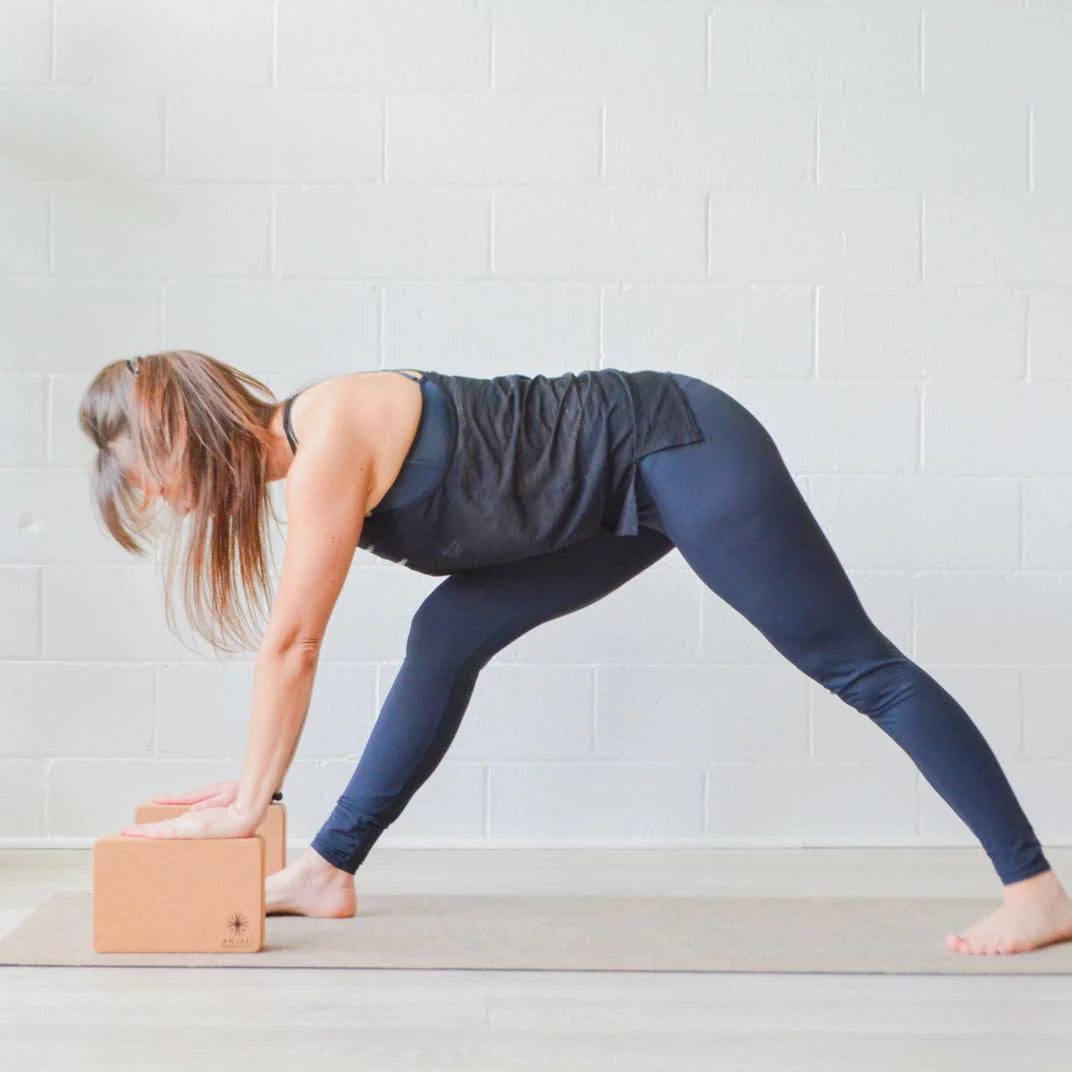 Woman practicing yoga with Anjali Generation cork yoga blocks on a light wooden floor against a white brick wall.