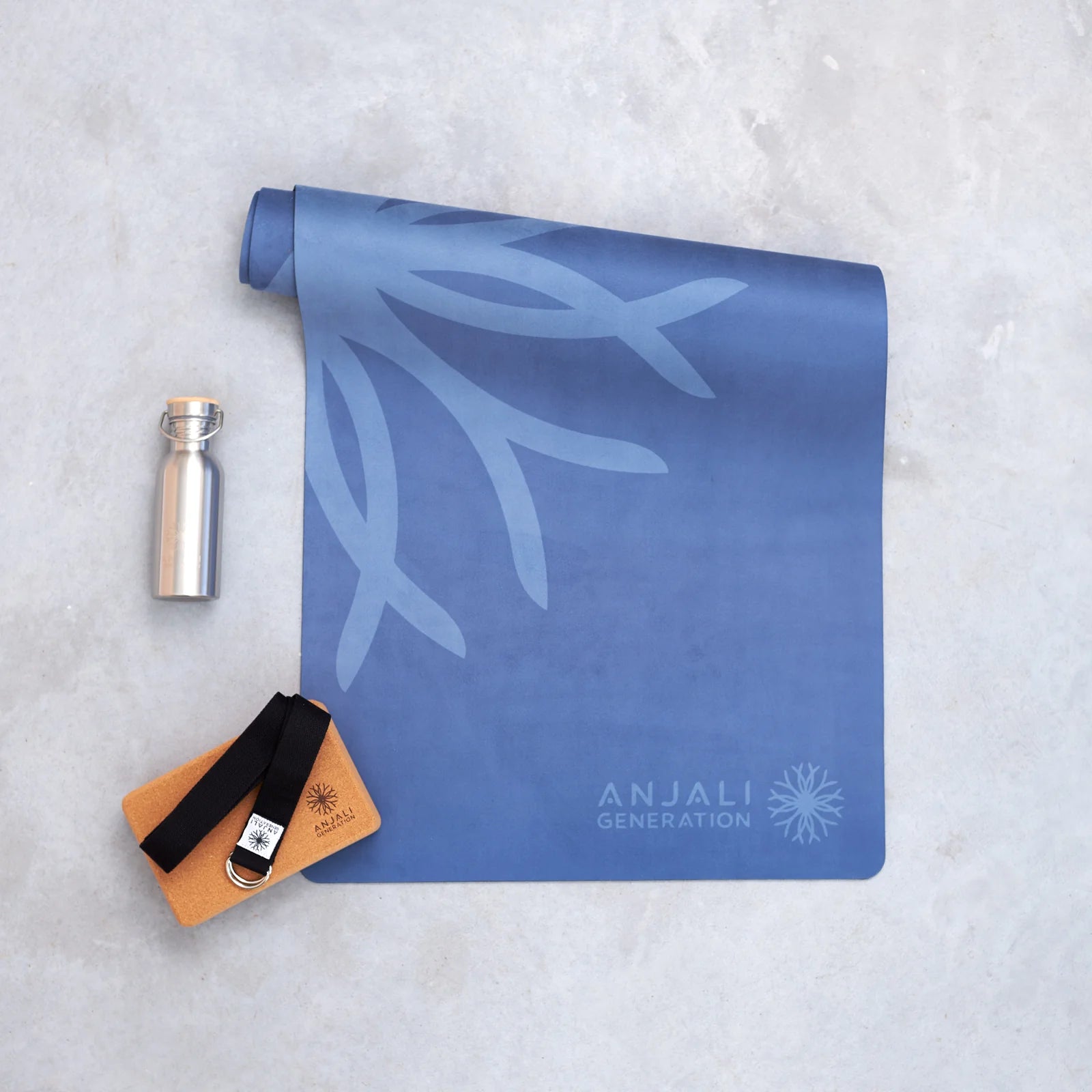 Blue yoga mat with 'Anjali Generation' branding, a silver water bottle, a black yoga strap and a cork yoga block on a light gray surface.