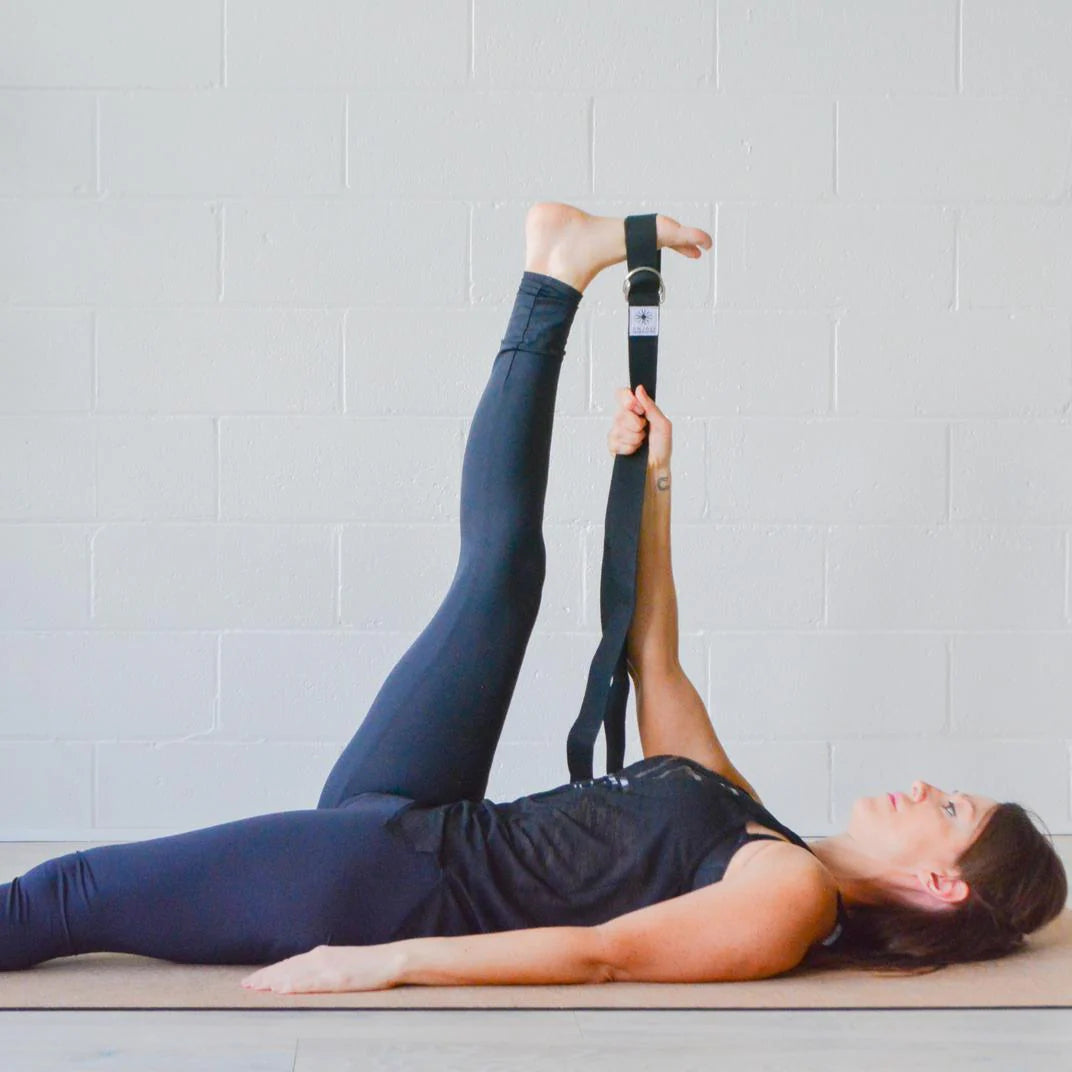 Person lying on a yoga mat with an Anjali Generation yoga strap around their legs against a light gray wall.