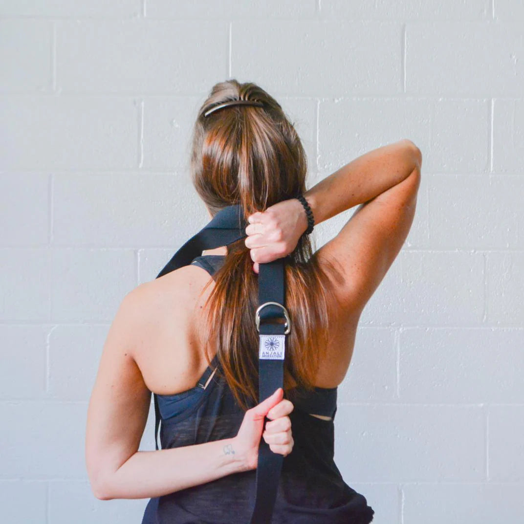 Woman using an Anjali Generation black yoga strap behind her back against a light gray wall