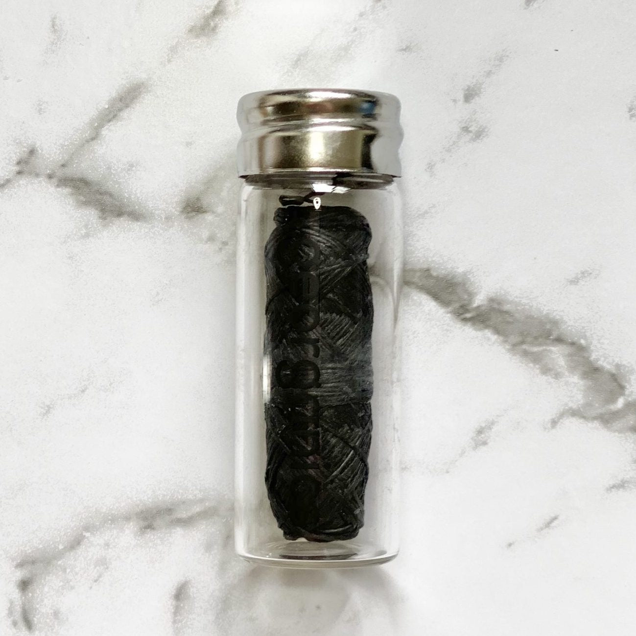 Clear glass container with a silver lid holding a black roll of Georganics activated charcoal natural floss on a marble background
