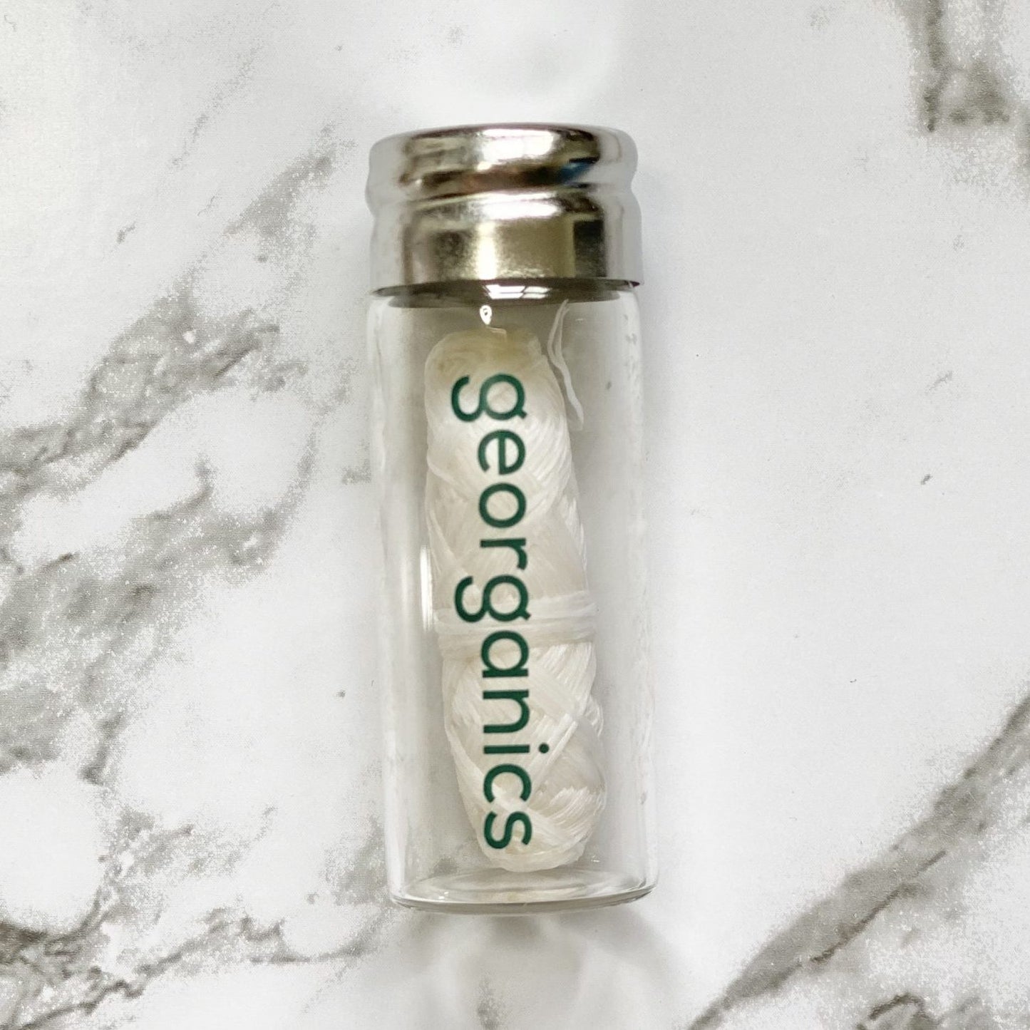 Clear glass container with a silver lid holding a white roll of Georganics spearmint natural floss on a marble background
