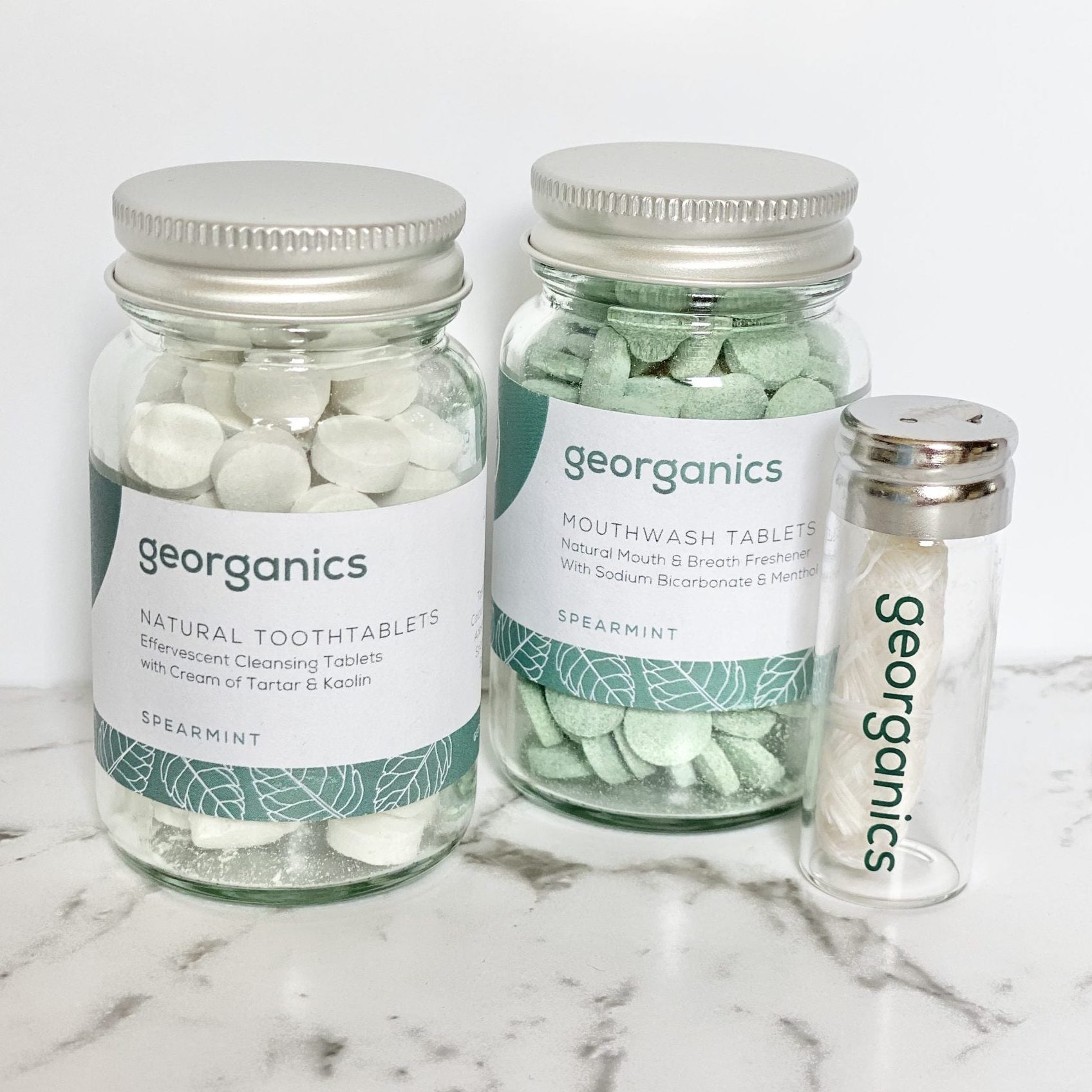 Three jars containing Georganics mouthwash tablets, toothtablets and natural floss on a marble surface.