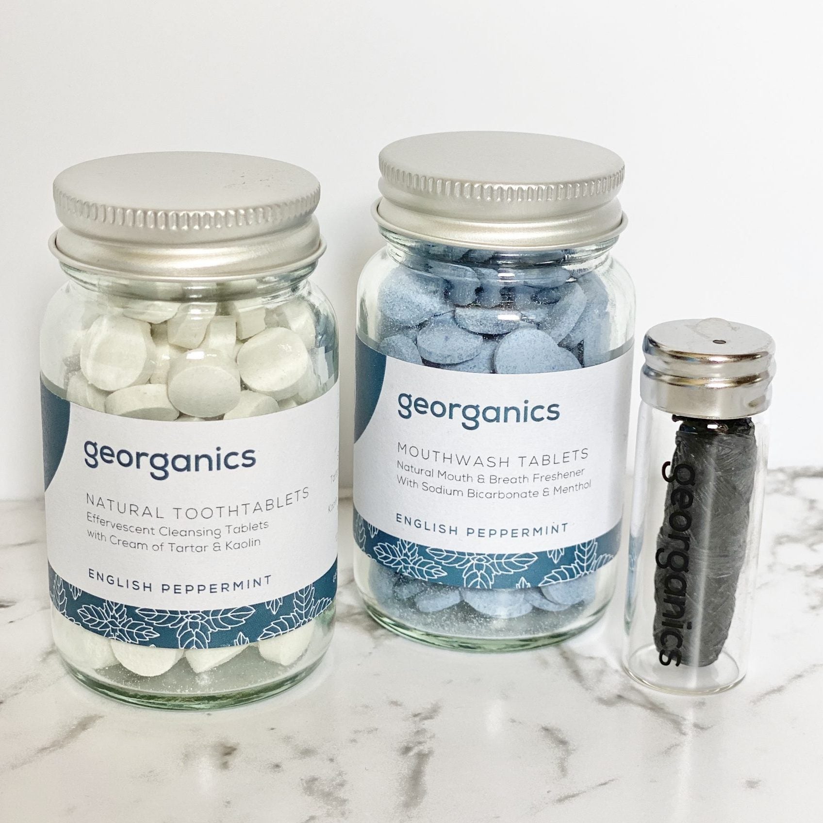 Three jars containing Georganics mouthwash tablets, toothtablets and natural floss on a marble surface.