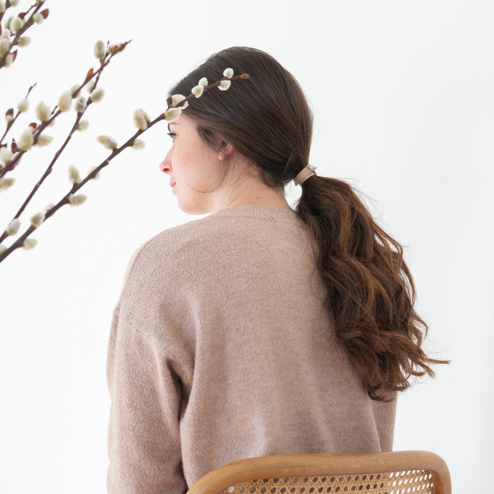 Woman with a ponytail using Kooshoo organic blond hair tie, wearing a beige sweater, sitting next to a willow branch with buds.