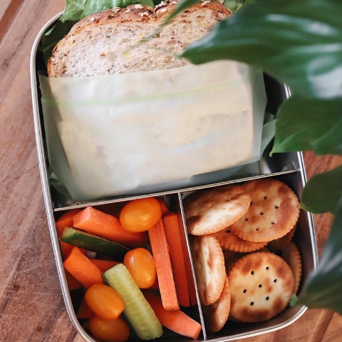 Lunchbox with a sandwich in a BioBag resealable compostable food bag, vegetables, and crackers on a wooden surface.