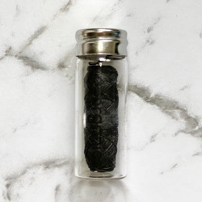 Clear glass container with a silver lid holding a black roll of Georganics activated charcoal natural floss on a marble background