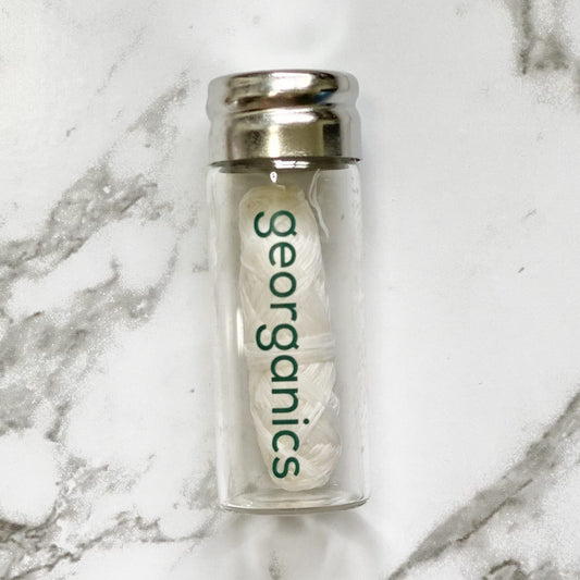 Clear glass container with a silver lid holding a white roll of Georganics spearmint natural floss on a marble background