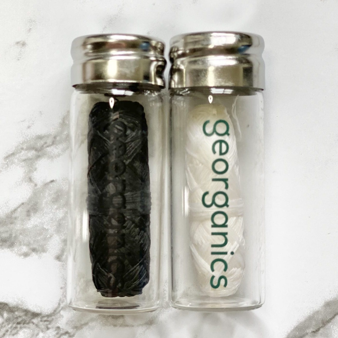 Two clear bottles with silver lids, one containing a black roll of Georganics activated charcoal natural floss, and the other containing a white roll of Georganics spearmint natural floss, on a marble surface.
