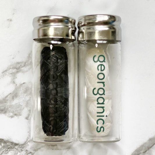 Two clear bottles with silver lids, one containing a black roll of Georganics activated charcoal natural floss, and the other containing a white roll of Georganics spearmint natural floss, on a marble surface.