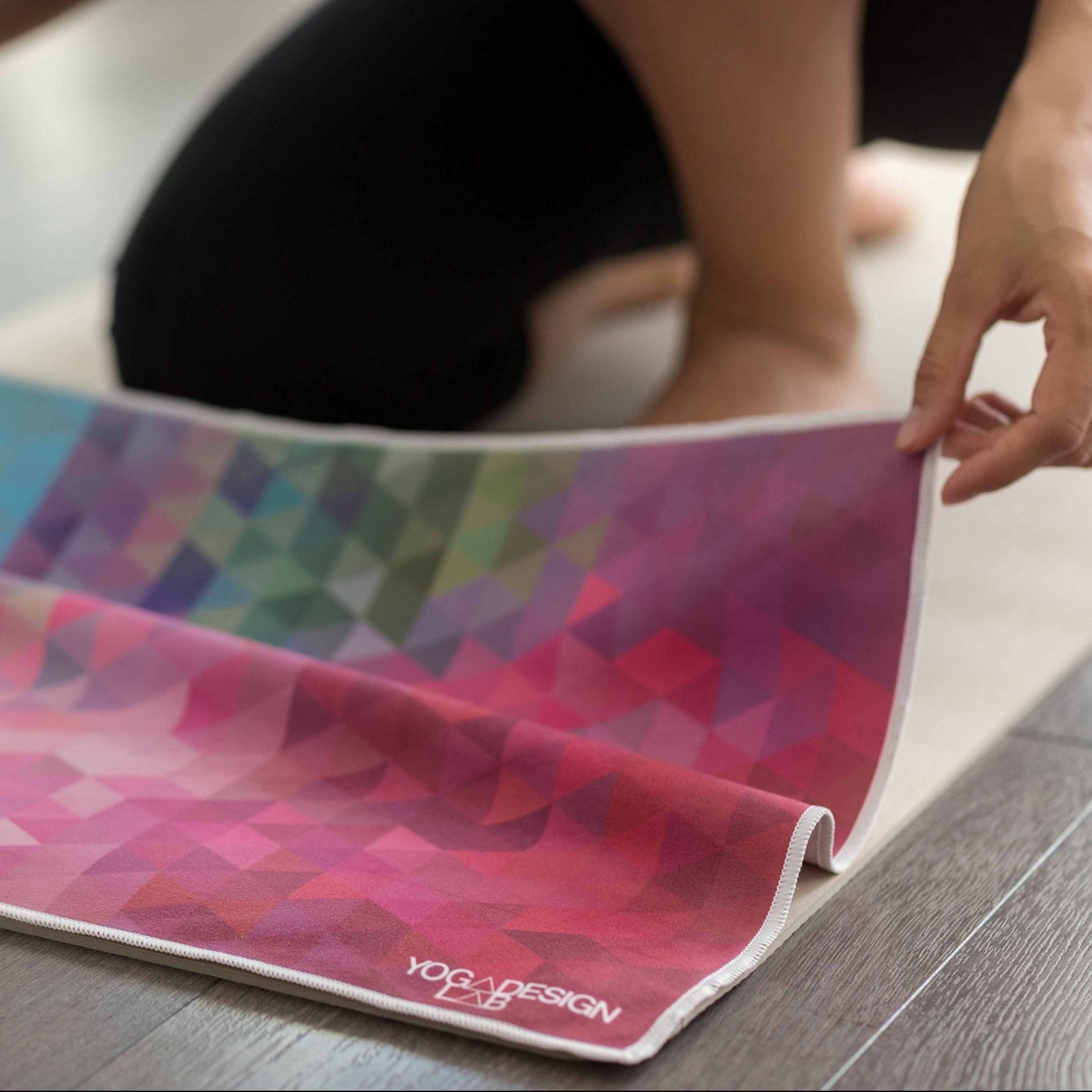 Person rolling up a colorful yoga hand towel with 'Yoga Design Lab' branding on a wooden floor.